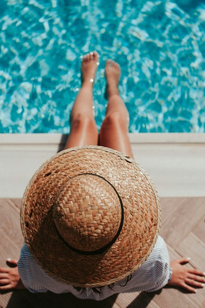 Straw Hat Woman wearing a straw hat sitting by a poolside with legs stretched out over turquoise water on a sunny day