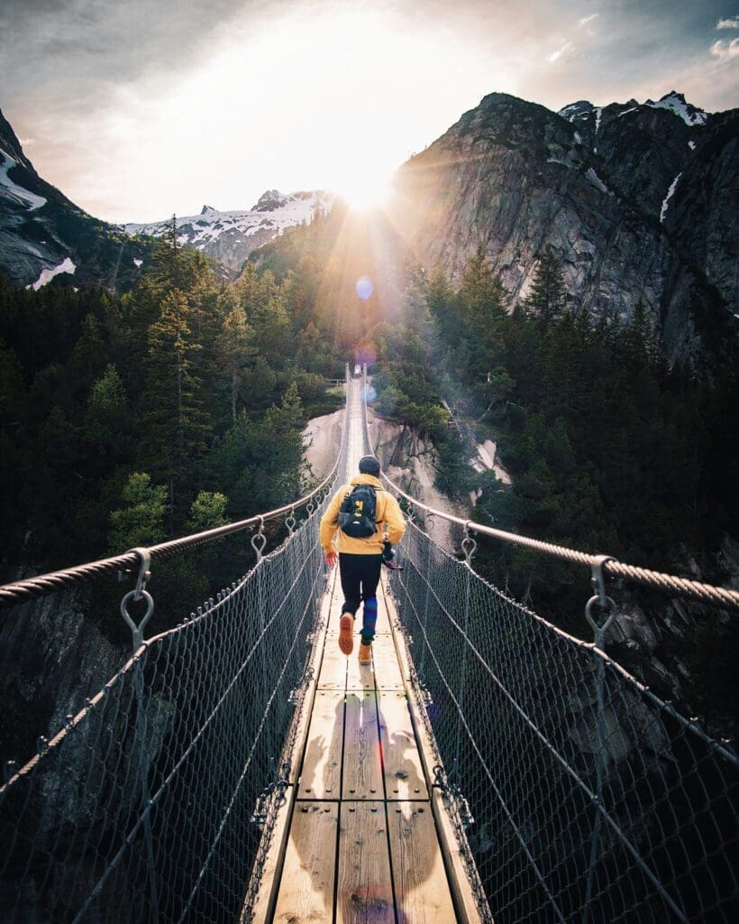 Photo by Fabio Comparelli on Unsplash Person walking across a suspension bridge in the mountains at sunrise, surrounded by pine trees and rocky peaks