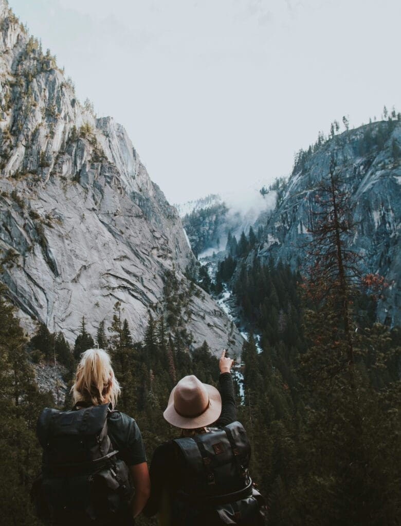 Two hikers with backpacks looking at a waterfall in a pine forest valley surrounded by dramatic rocky cliffs and mist in the mountains