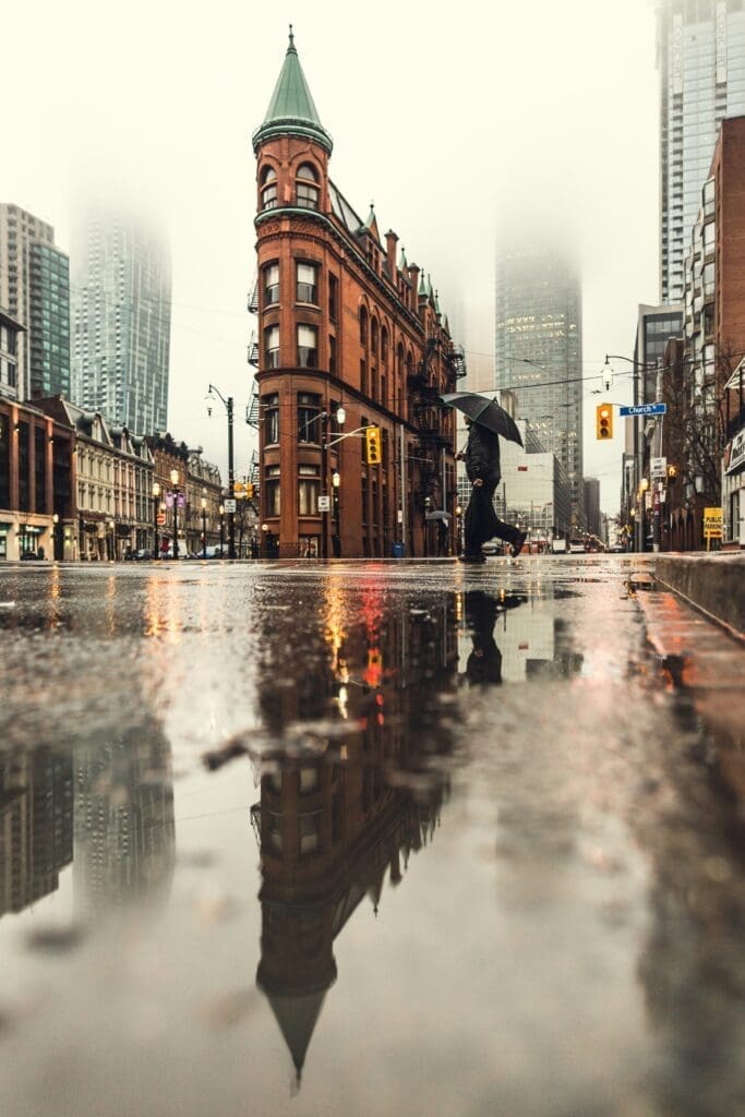 Gooderham Building Person with umbrella crossing a rainy street in front of the Flatiron-style red brick Gooderham Building in downtown Toronto with reflections in puddles and foggy skyscrapers in the background