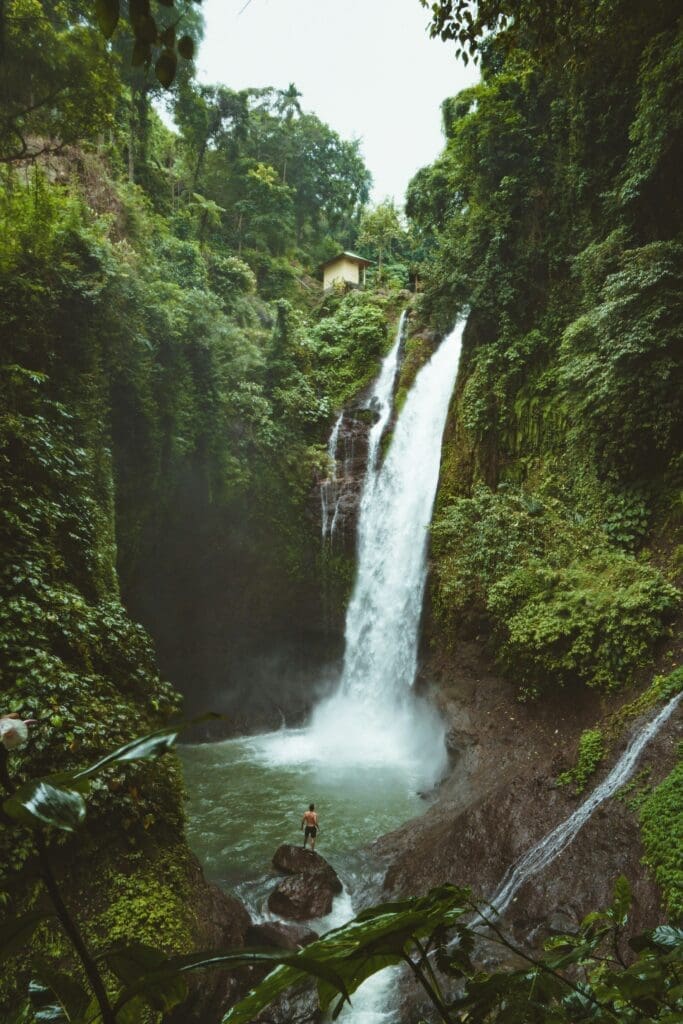 Waterfall Man standing on a rock at the base of a tall jungle waterfall surrounded by lush green rainforest