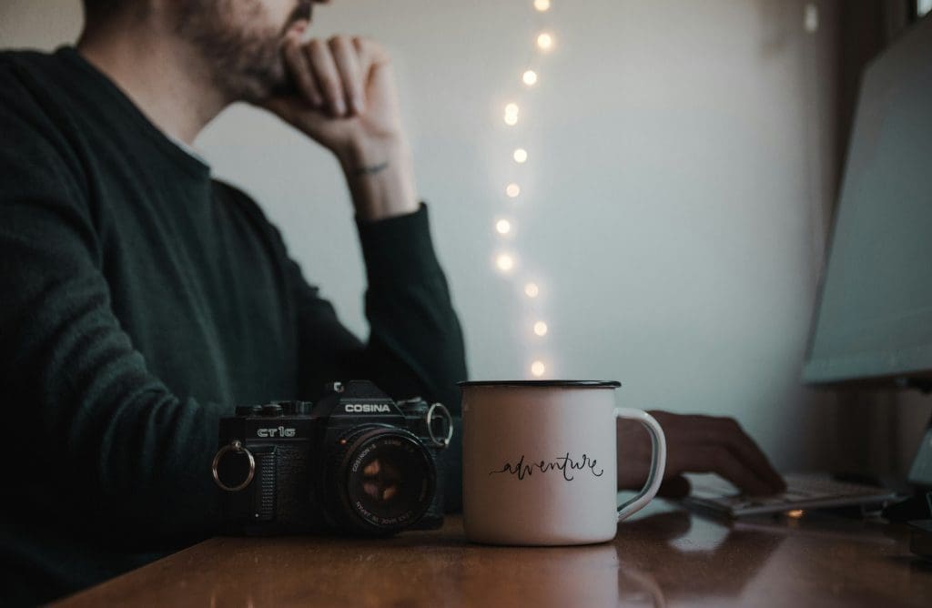 Photo by Sergio Rodriguez Portugues Del Olmo on Unsplash Man working at desk with vintage Cosina camera and coffee mug labelled adventure