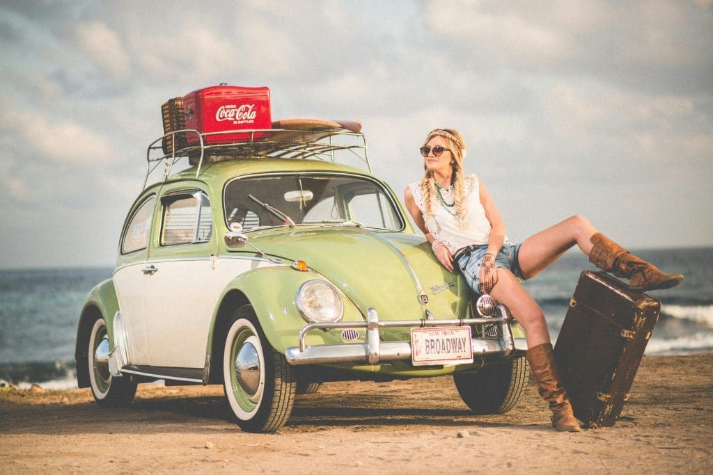 Photo by amos bar zeev on unsplash Woman in sunglasses and boots leaning on a vintage green Volkswagen Beetle with luggage and a red Coca-Cola cooler on the roof, parked by the seaside