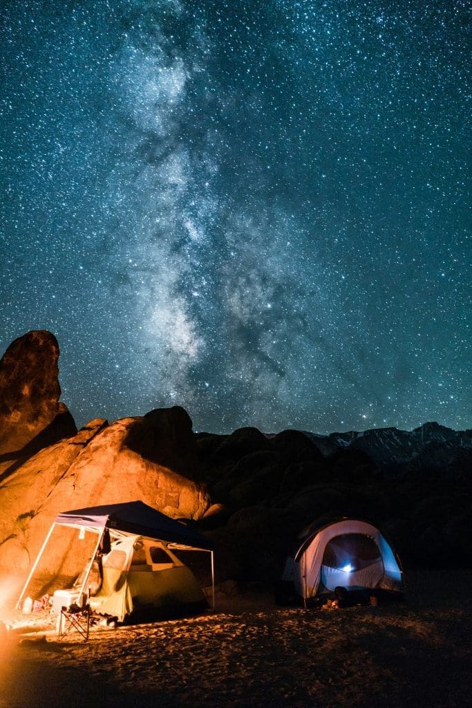 Camping tents and campfire under a clear night sky filled with stars and the Milky Way above rocky desert mountains