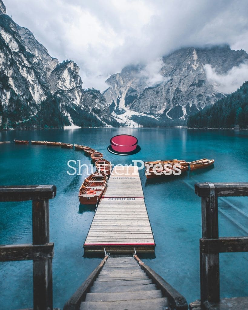 Wooden pier with rowboats on turquoise alpine lake surrounded by dramatic mountains and mist, ShutterPress watermark