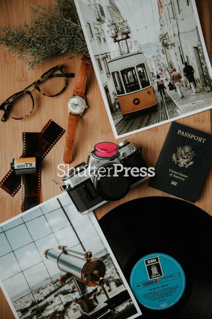 Flat lay of vintage travel and photography items including a Canon film camera, watch, glasses, passport, vinyl record and Lisbon tram photo, ShutterPress watermark