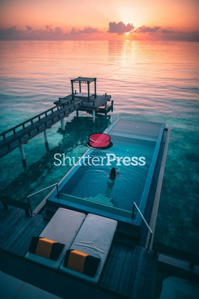 Infinity pool over the ocean at sunset with woman swimming, wooden pier and tropical seascape, ShutterPress watermark