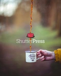 Coffee pouring into enamel camping mug with The Adventure Begins design, outdoors in nature, ShutterPress watermark