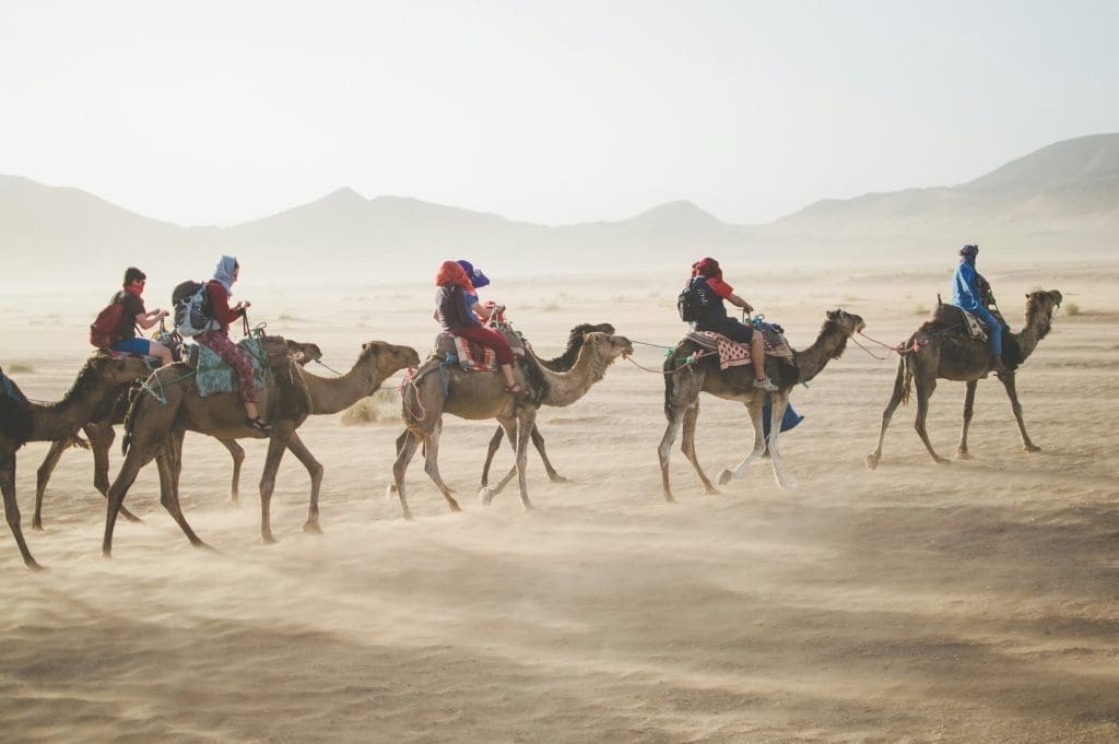 Photo by vaida tamosauskaite on unsplash Group of travellers riding camels across a desert with mountains in the background, Morocco Sahara tour