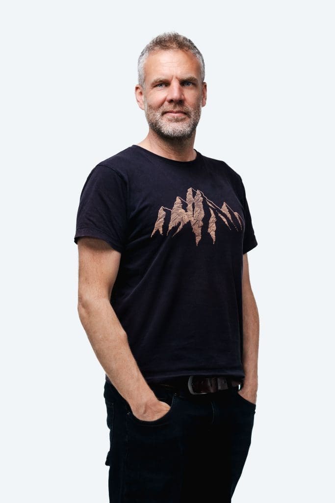 Portrait of Olly Bowman with short grey hair and beard, wearing a black t-shirt with a mountain design, neutral background.