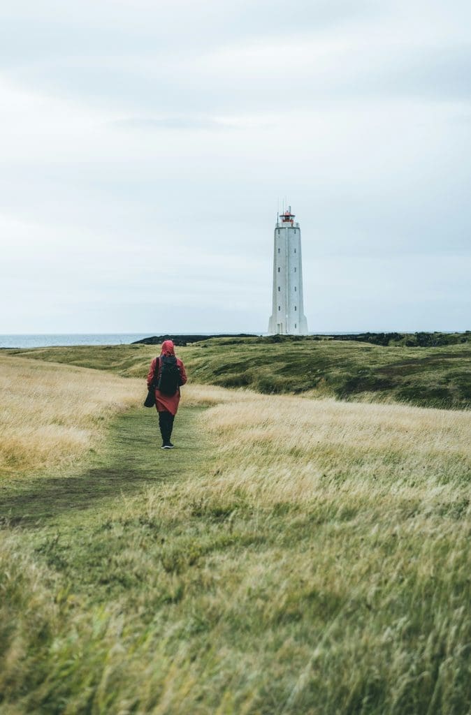 Photo by Alexander Milo on Unsplash Person in red coat walking across grassy coastal path towards tall white lighthouse under grey sky
