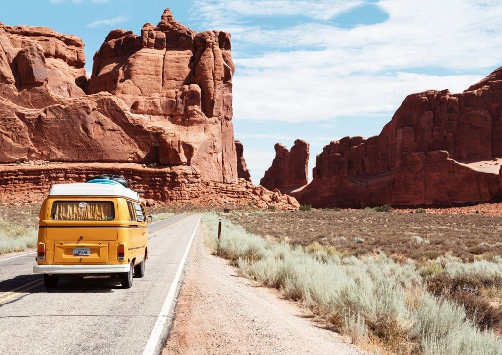 Photo by Dino Reichmuth on Unsplash Yellow camper van with ‘EXPLORE’ number plate driving down a deserted road through red sandstone cliffs and desert scrub under a partly cloudy sky.