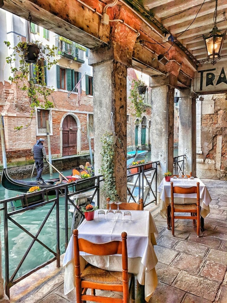 Photo by Igor Oliyarnik on Unsplash Gondolier paddling past tables of a waterside café under rustic portico in Venice canal