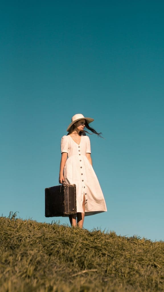 Photo by Iurii Melentsov on Unsplash Woman in white dress and sun hat standing on grassy hill holding vintage suitcase under clear blue sky