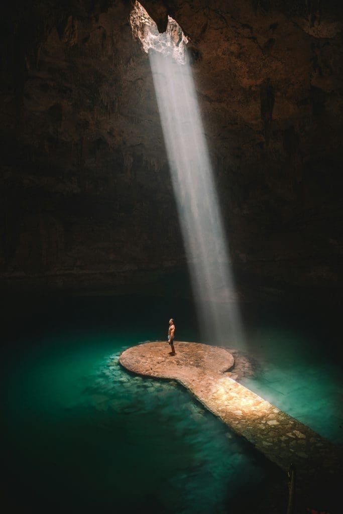 Photo by Jared Rice on Unsplash Man standing on stone platform in cenote cave with sunbeam streaming through roof onto turquoise water