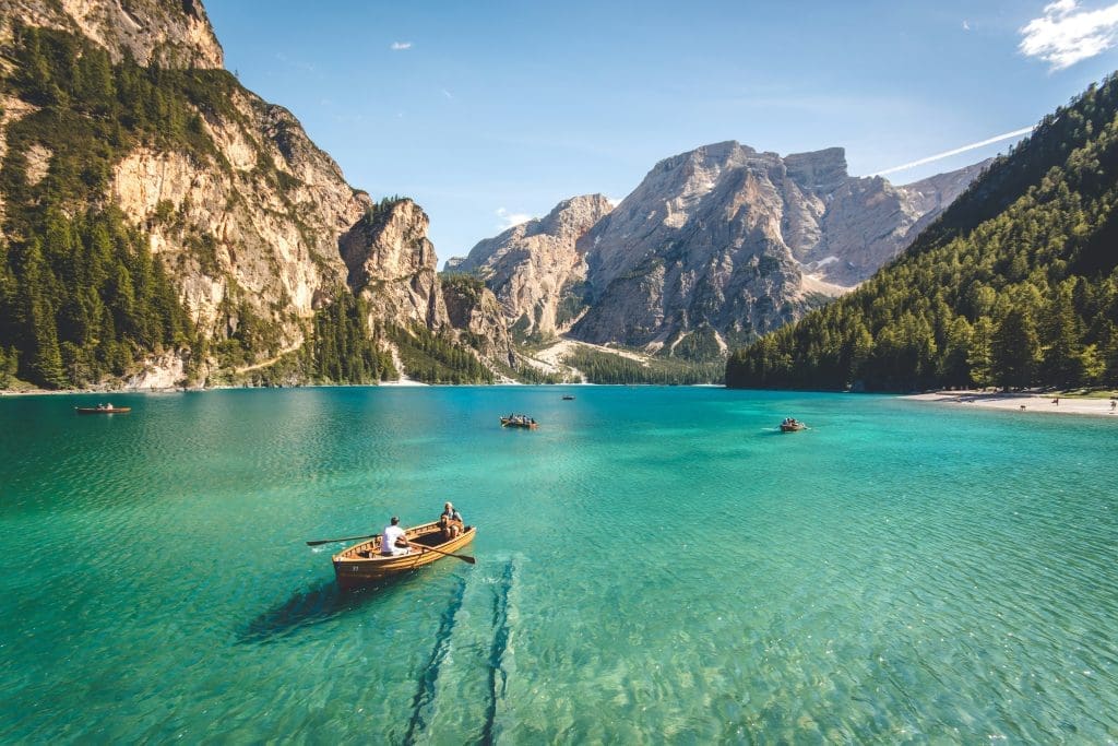 Photo by Pietro De Grandi on Unsplash Turquoise alpine lake surrounded by rugged forested peaks, wooden row-boats with people move gently across the clear water under a bright sky.