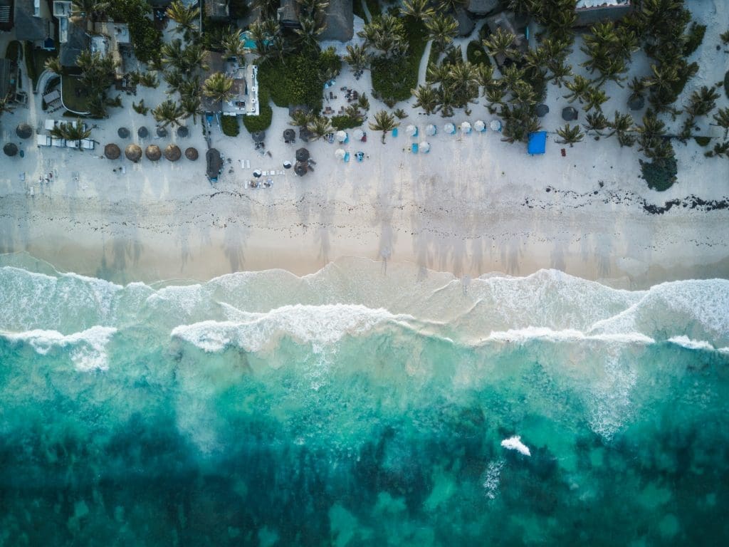 Photo by Spencer Watson on Unsplash Aerial view of turquoise waves washing onto white sandy beach lined with palm trees and umbrellas in tropical resort